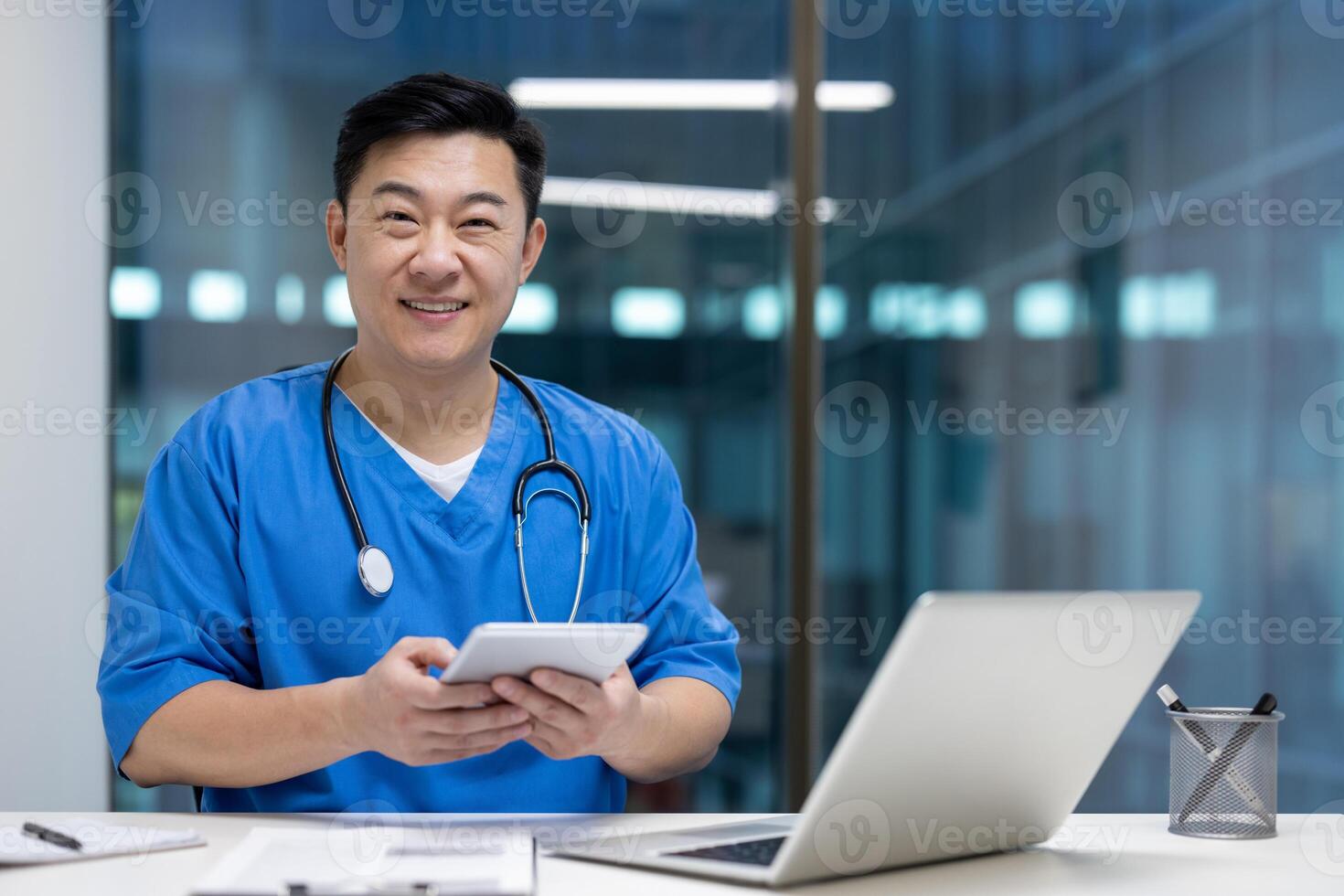 Asian man doctor holds tablet smiling in modern office with laptop, representing technology in healthcare. Blue medical uniform, stethoscope around neck, creating professional atmosphere. photo