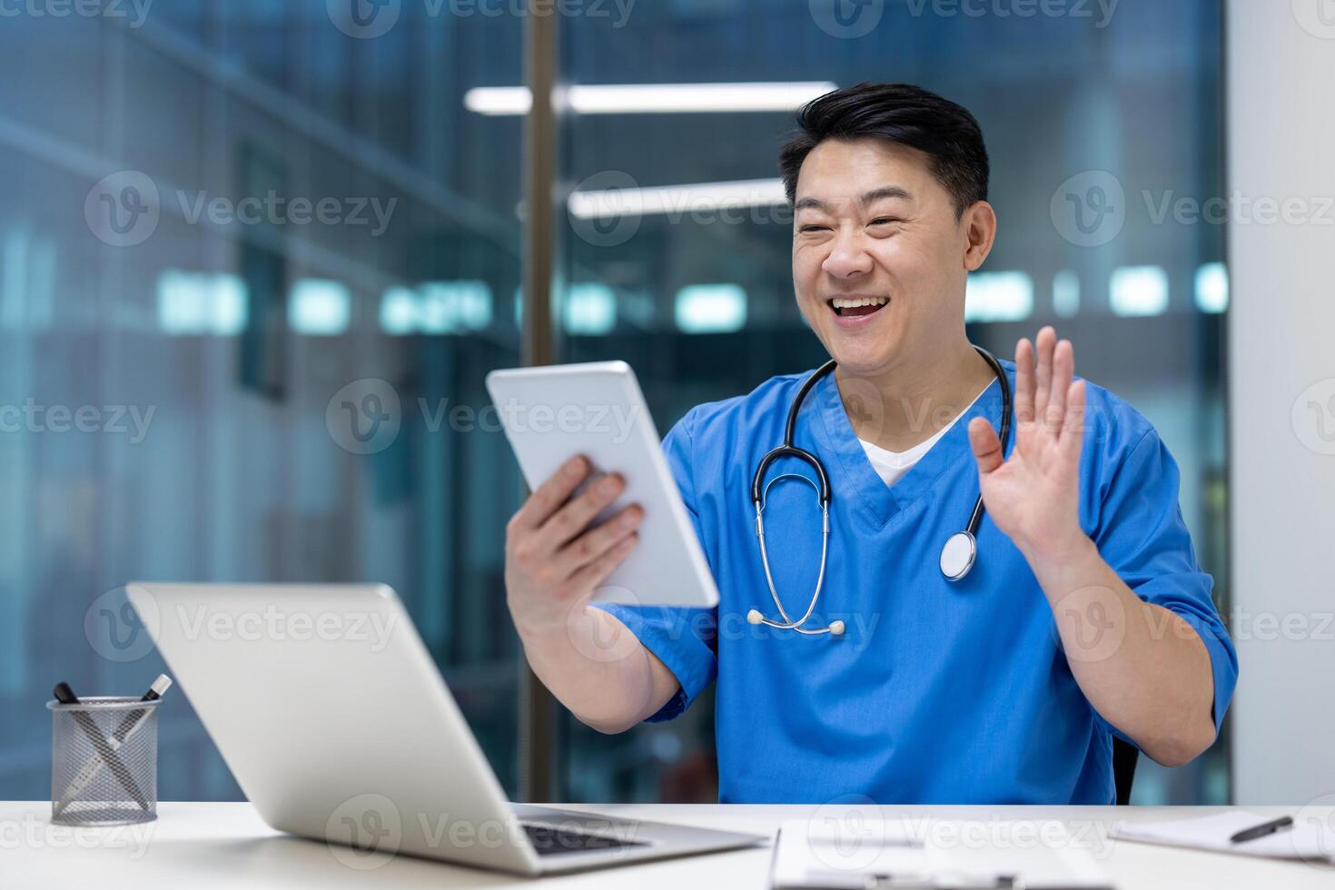 Asian man doctor in blue scrubs engaging in call on tablet, smiling and waving, showcasing communication and technology in healthcare, laptop and phone visible on desk. photo