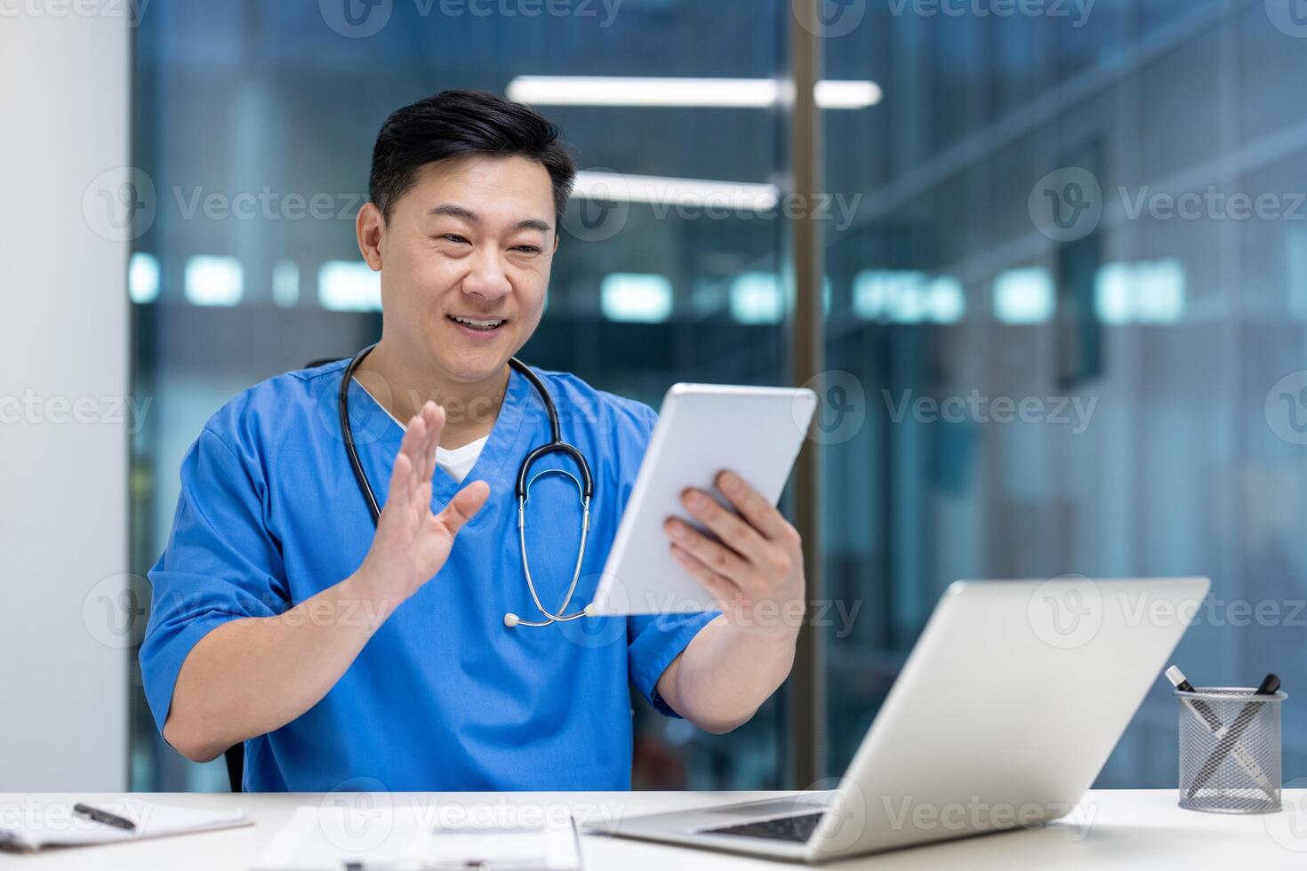Asian man doctor in blue scrubs using tablet for virtual consultation in office. Engaged in call, smiling confidently, with laptop on desk, conveying professionalism and technology. photo