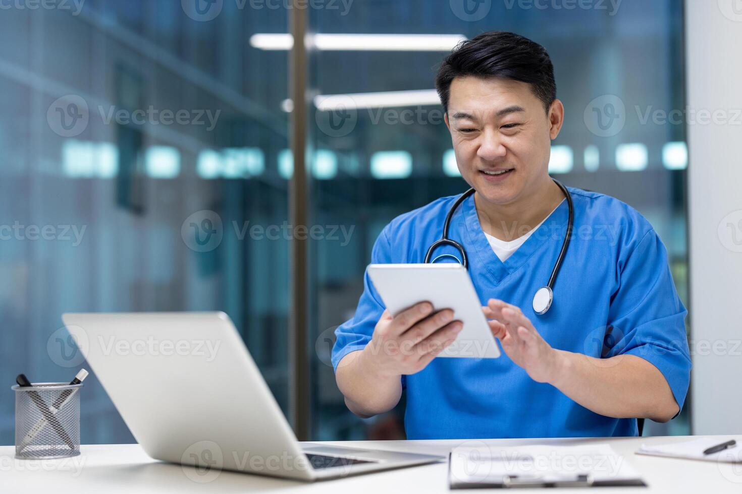 Asian man doctor using tablet and laptop in office, showcasing efficiency and modern healthcare solutions. Wearing scrubs, smiling, focused on digital screen, enhancing medical productivity. photo