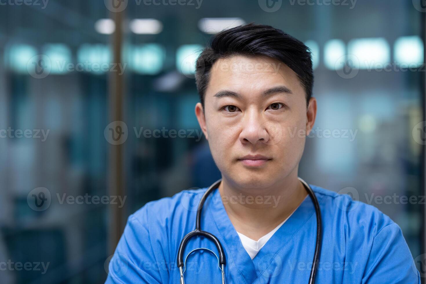 Asian man doctor in blue scrubs stands confidently with stethoscope in modern healthcare facility. Expression serious, projecting professionalism and dedication in medical field. photo