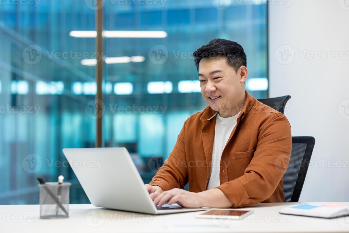 Asian businessman using laptop in bright modern office. Smiling while working, wearing casual shirt. Represents concepts of productivity, technology, teamwork, and professionalism. Includes phone. photo