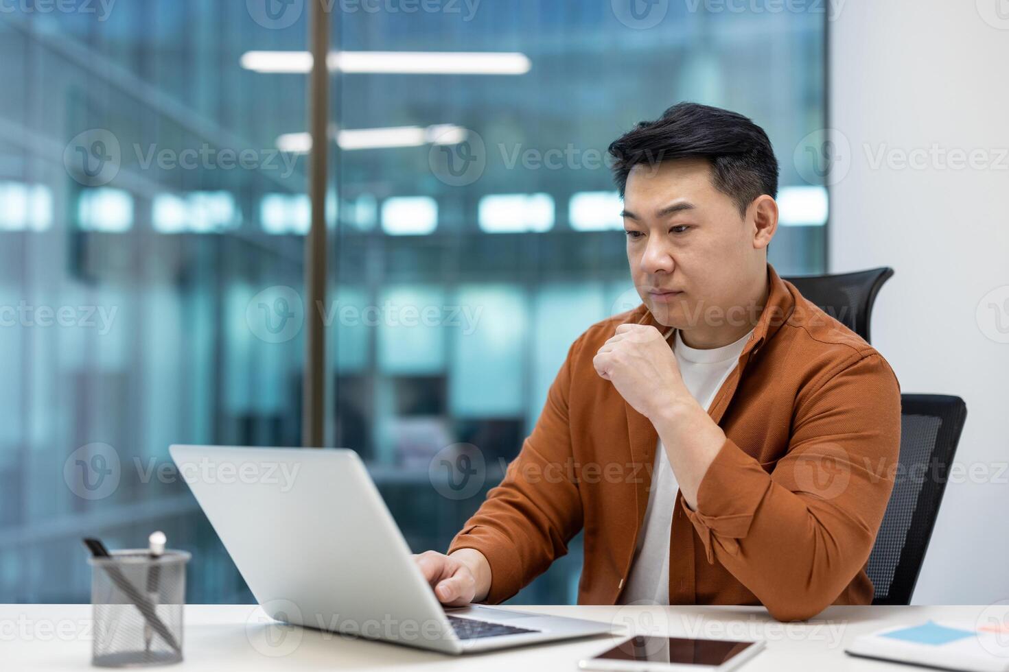 Asian man businessman working in modern office, focused on laptop. Casual attire signifies balance between professionalism and comfort, surrounded by large windows enhancing open workspace. photo