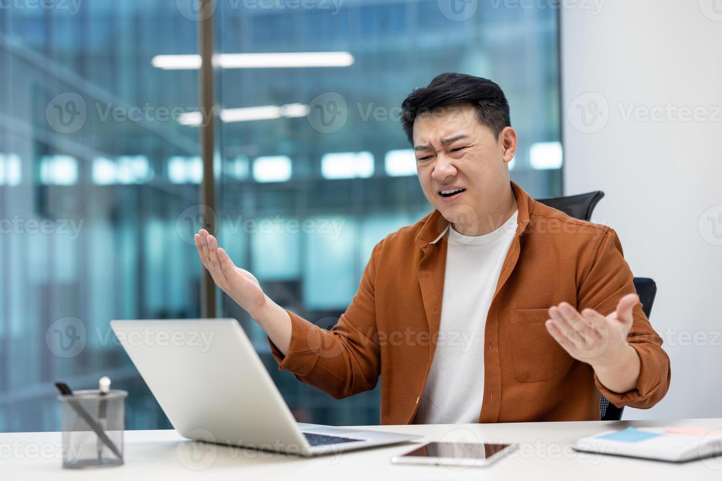 Asian man businessman showing frustration while working on laptop in office. Expressing stress and confusion with hands up and bewildered expression, phone on desk. photo