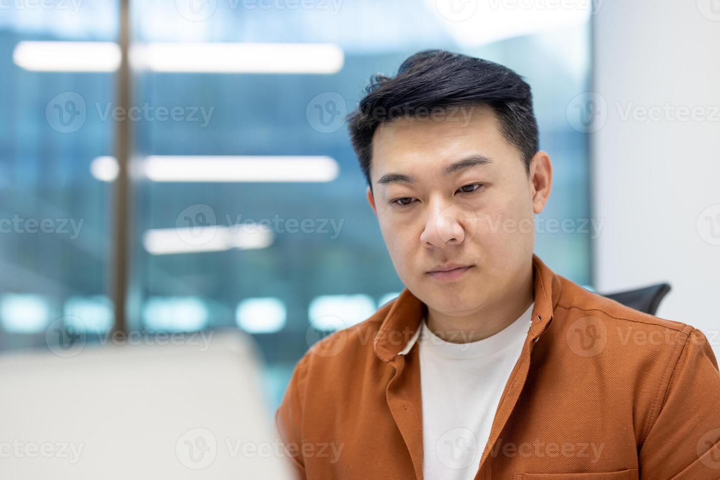 Asian man businessman at desk in modern office setting wearing orange jacket focused on work. Large glass windows create bright atmosphere. Represents professionalism and dedication. photo