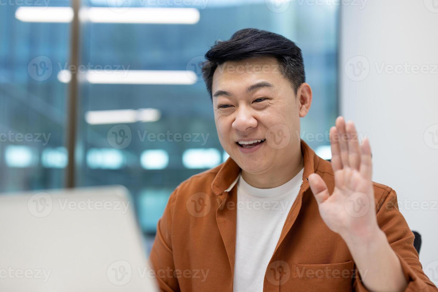 Asian man businessman engaging in virtual meeting, smiling, waving at laptop screen in casual outfit. Modern office setting, focus on communication and technology. photo