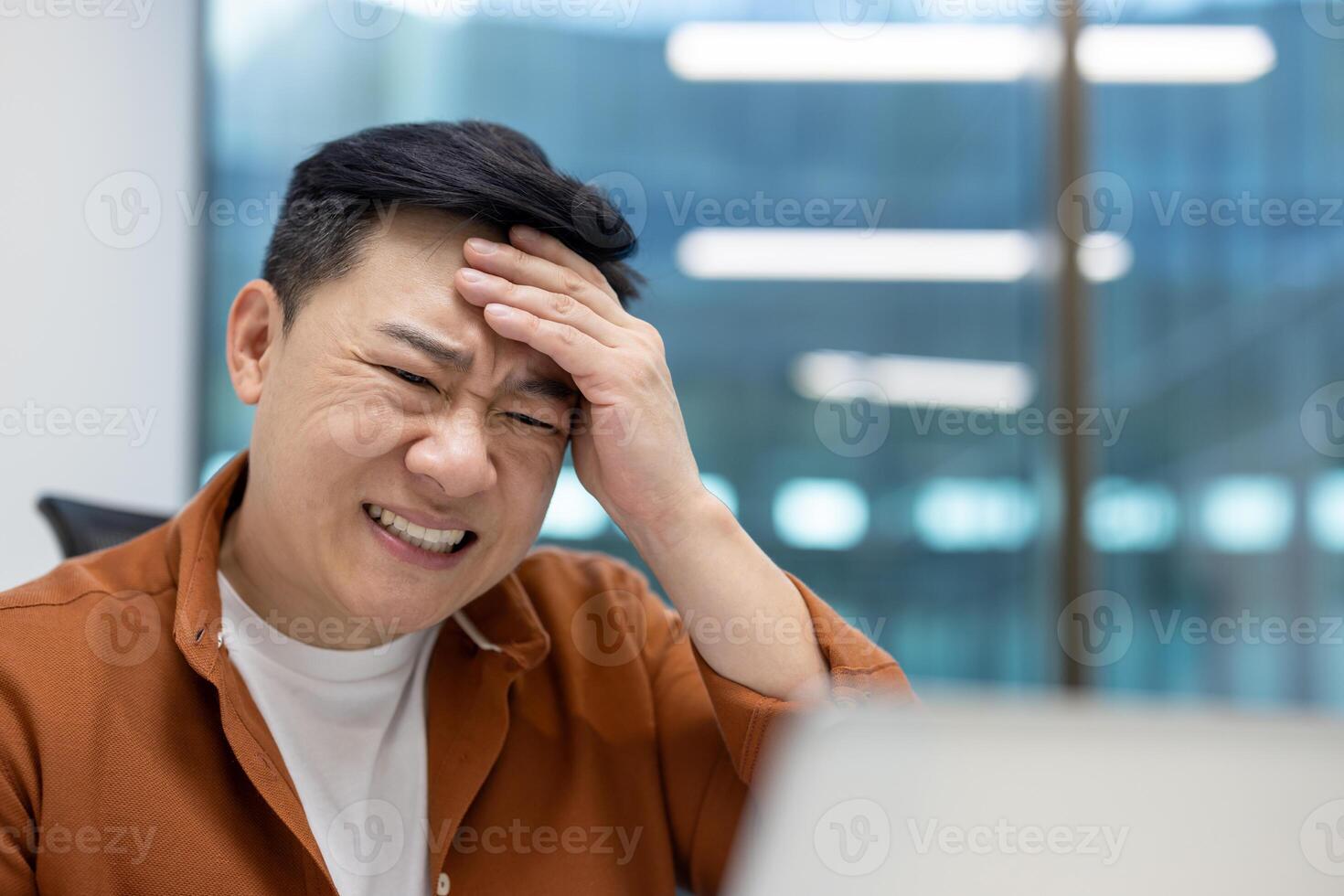 Asian man businessman feeling stress at work with hand on forehead, expressing frustration and fatigue in modern office setting. Business stress and overwhelmed emotions visible. photo