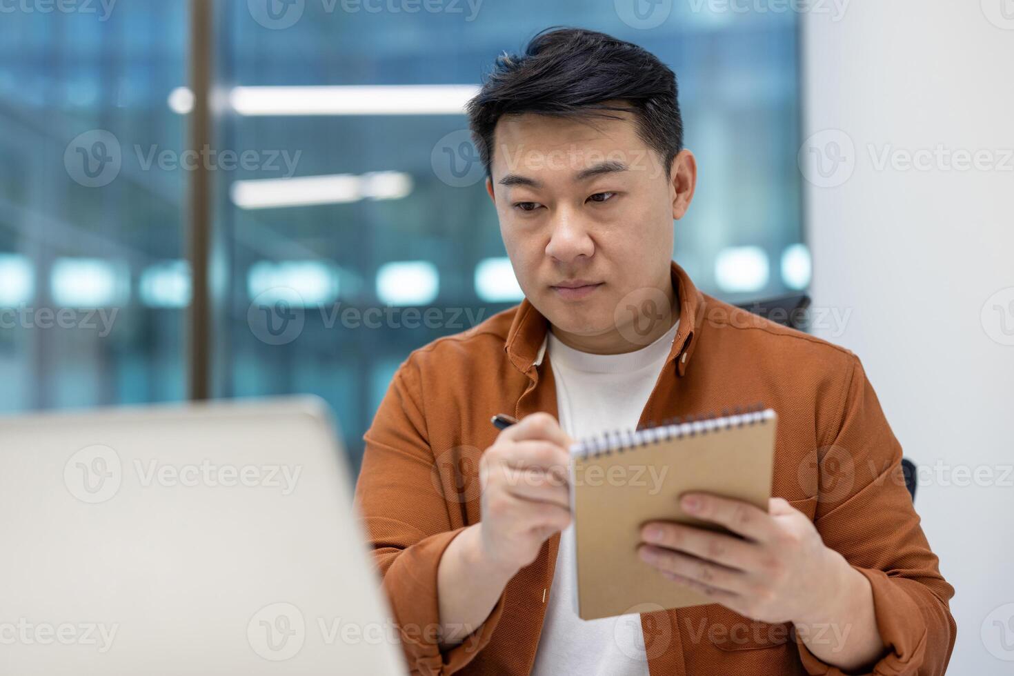 Asian man businessman taking notes on call meeting with laptop. Focused on work, wearing casual attire, modern office setting. Technology and communication concept. photo
