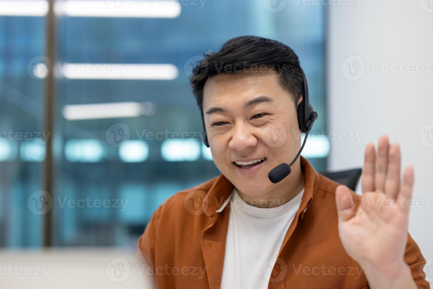 Asian man businessman in call using headset for online meeting in modern office. Smiling, waving hand, showcasing remote communication, teamwork, technology for workplace connectivity. photo