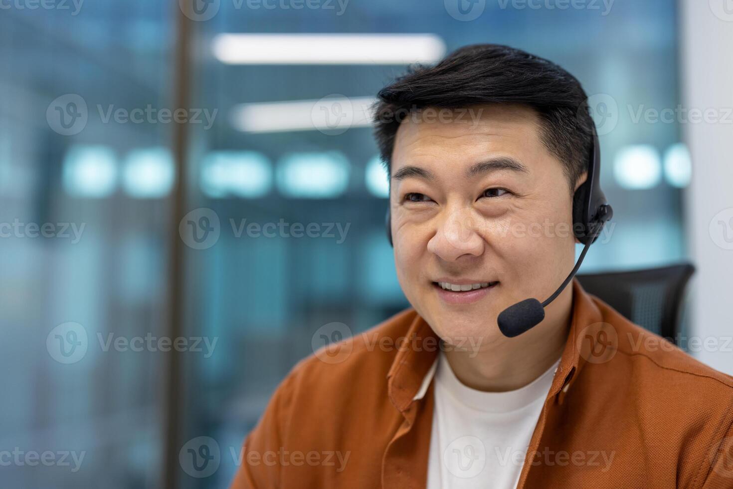 Asian man businessman smiling at desk in modern office, wearing headset for online communication. Bright, focused expression suggests positivity, effective engagement in digital business meeting. photo