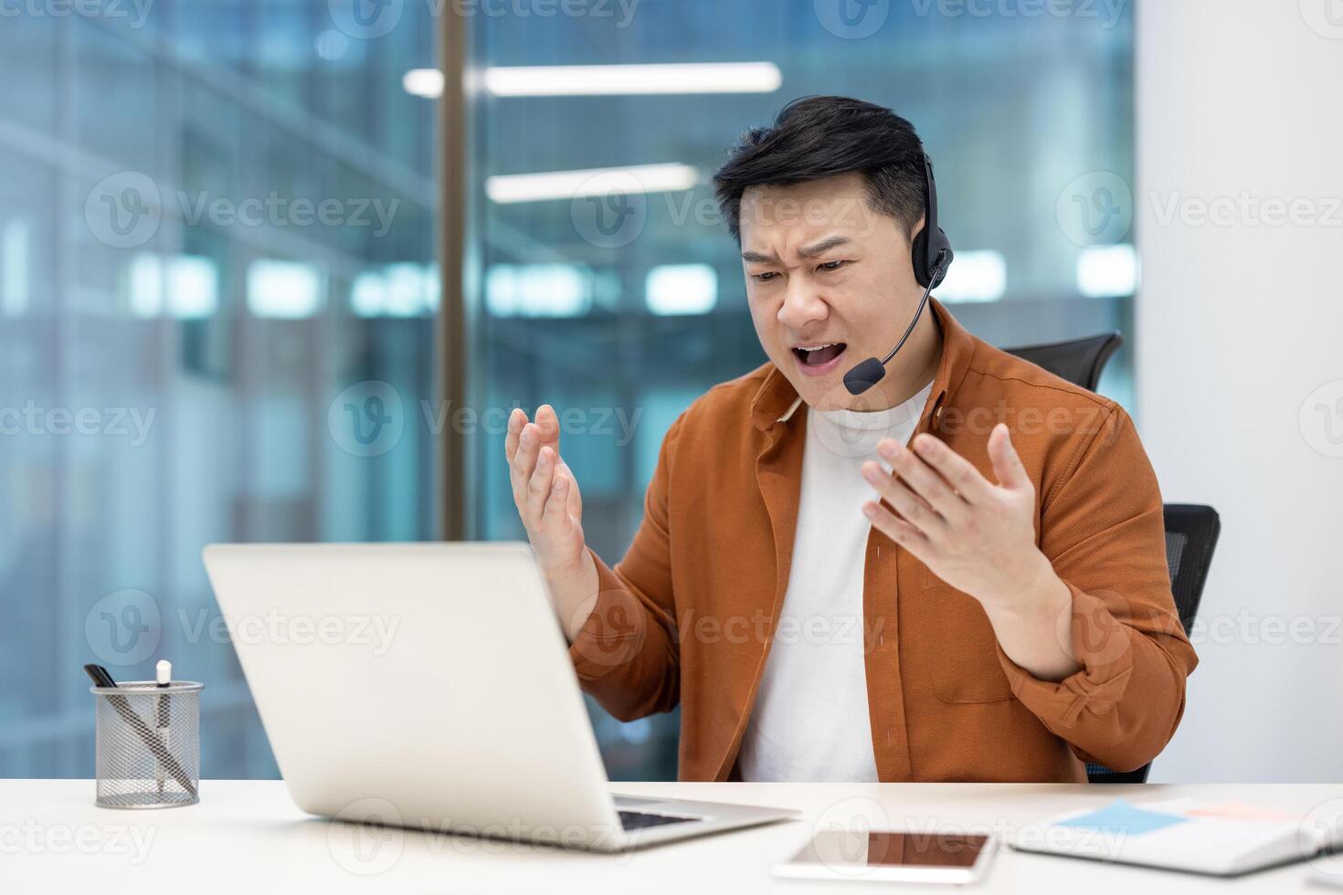 Angry man with headset talking while sitting at laptop inside office, dissatisfied employee yelling at interlocutor, unable to communicate with client. photo