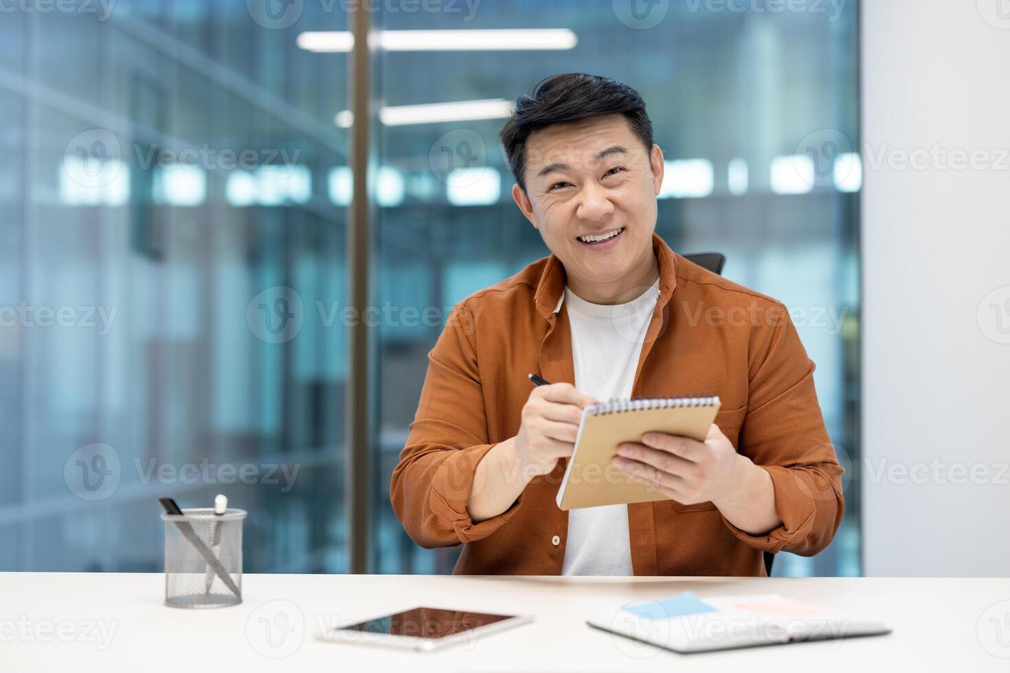 Asian man with notebook smiling and looking at camera, online meeting and business conference remotely. Businessman at workplace inside office at workplace. photo