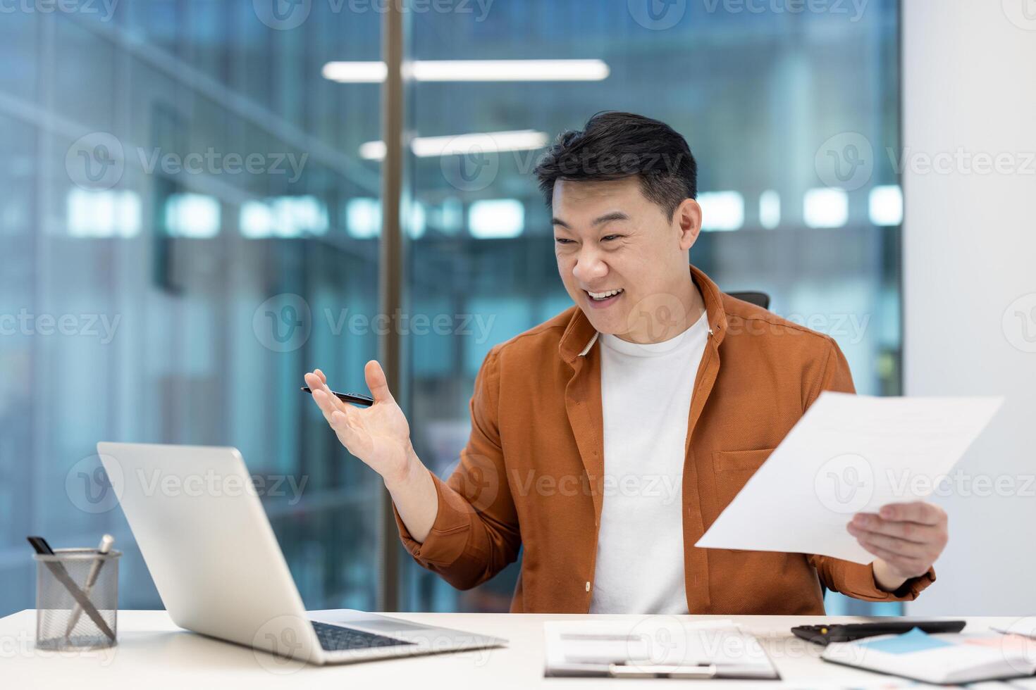 Asian man mature businessman interacting online, using a laptop, holding papers, and smiling in an office, conveying professionalism, planning, and communication during a remote business presentation photo