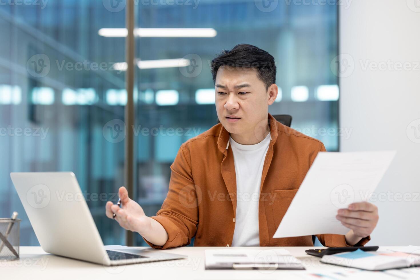 Asian man examining documents with a critical expression while using a laptop in office, reflecting a moment of professional questioning. photo