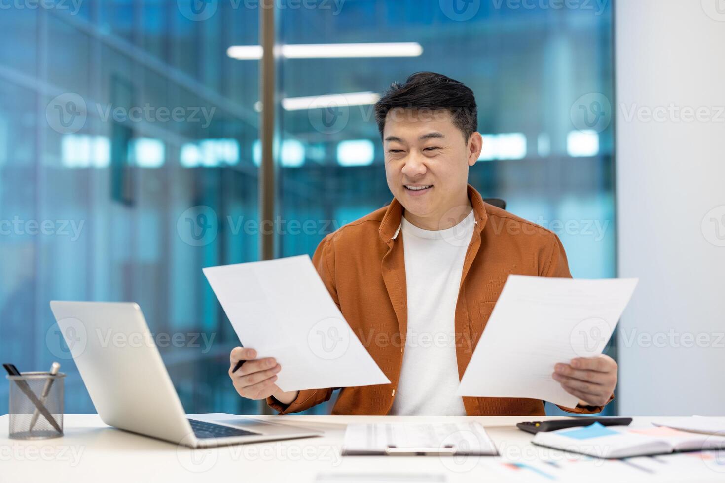 Smiling Asian businessman in casual attire reviewing documents at desk with laptop, symbolizing focus and satisfaction. Ideal for business themes, entrepreneurship, motivation. photo