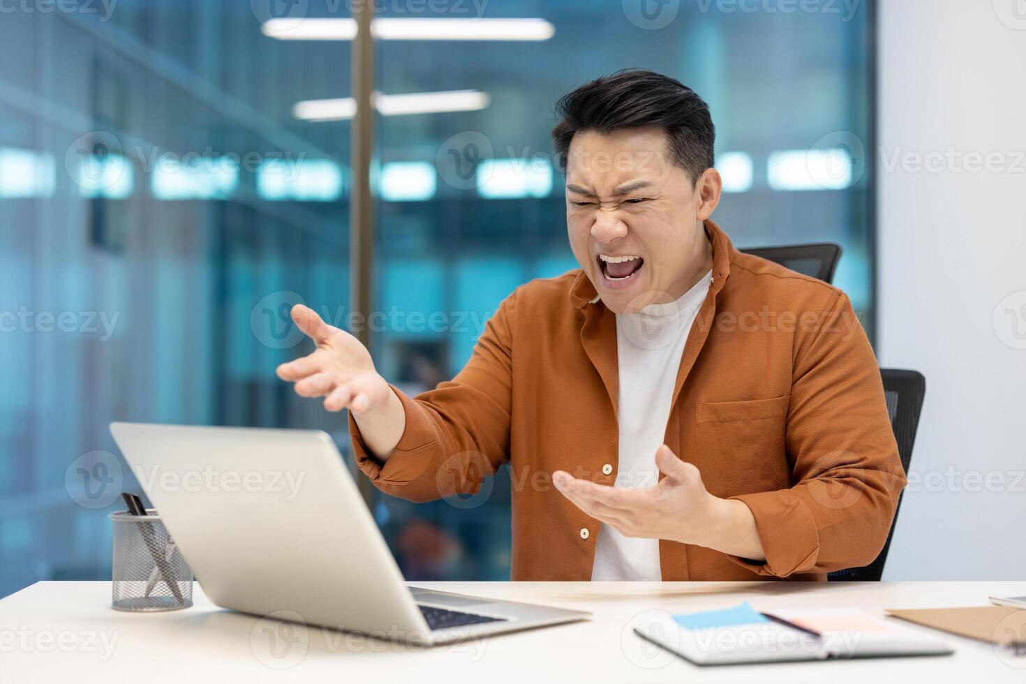 Asian businessman feeling frustrated while working on laptop. Expressing anger in a modern office setting. Concepts of stress, work pressure, emotions, and business challenges. photo