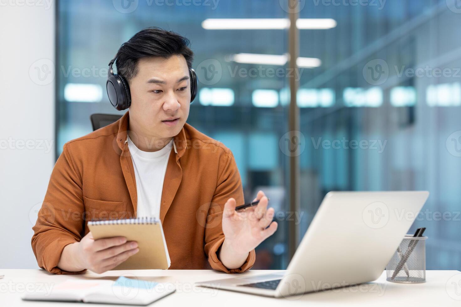 Asian man wearing headphones using a laptop for an online meeting, dressed casually in an office setting. He holds a notepad in one hand and writes with a pen, showing focus and productivity. photo