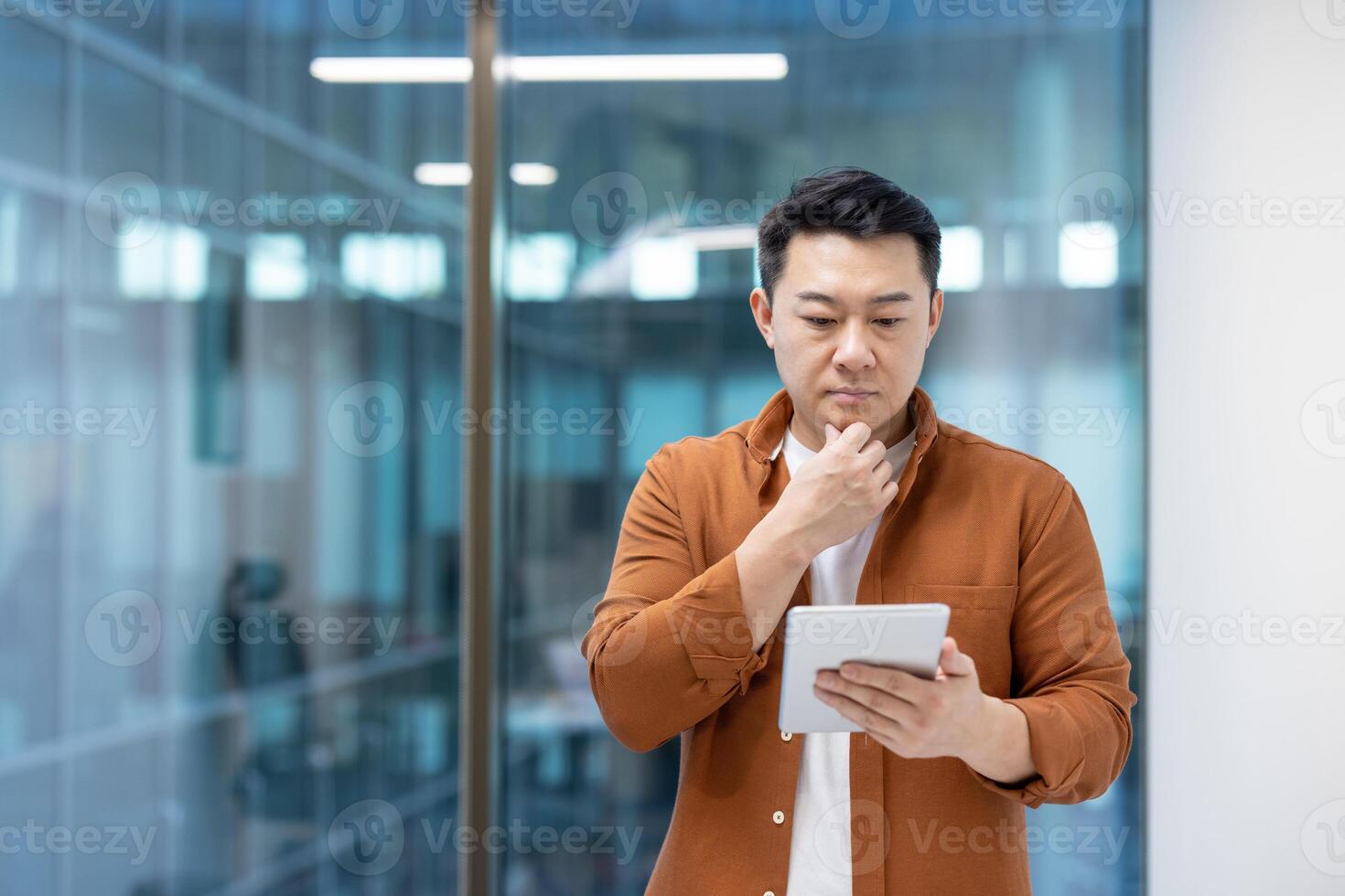 Asian man focusing on a task using a digital tablet in an office. His thoughtful expression suggests active problem-solving, concentration, and workplace productivity within a professional setting. photo