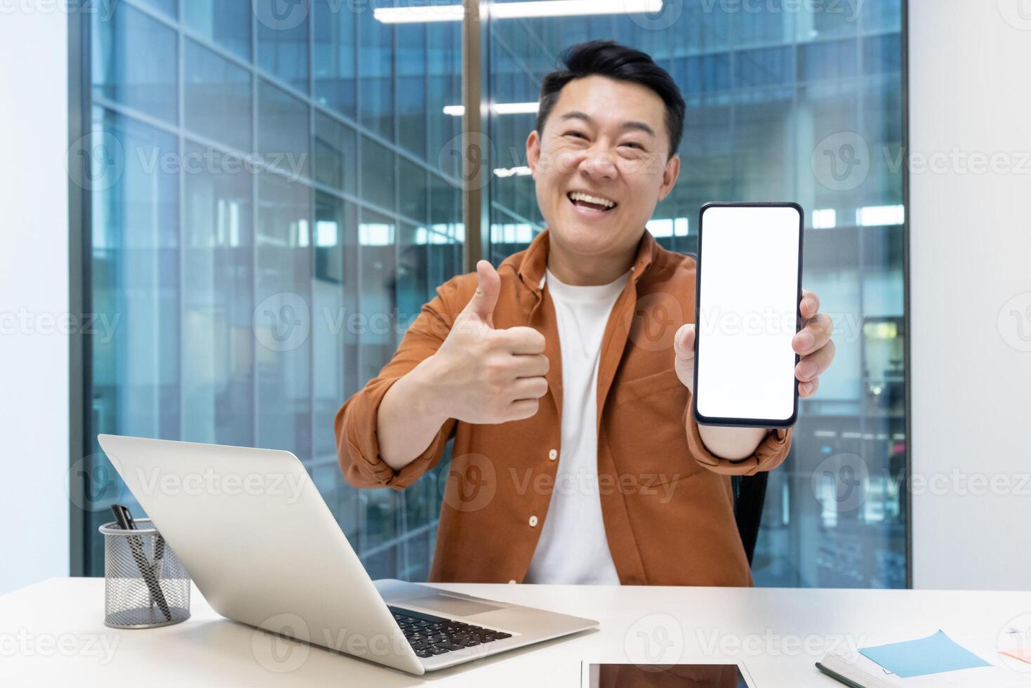 Asian man presenting and showing a smartphone while sitting at a desk in an office. The individual appears confident and joyful, highlighting the blank screen for any customizable display photo