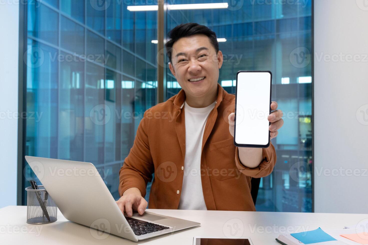 Asian businessman in office holds smartphone with blank screen, smiles confidently. Works on laptop at desk, displaying technology professionalism, innovative spirit. Modern workspace vibes. photo