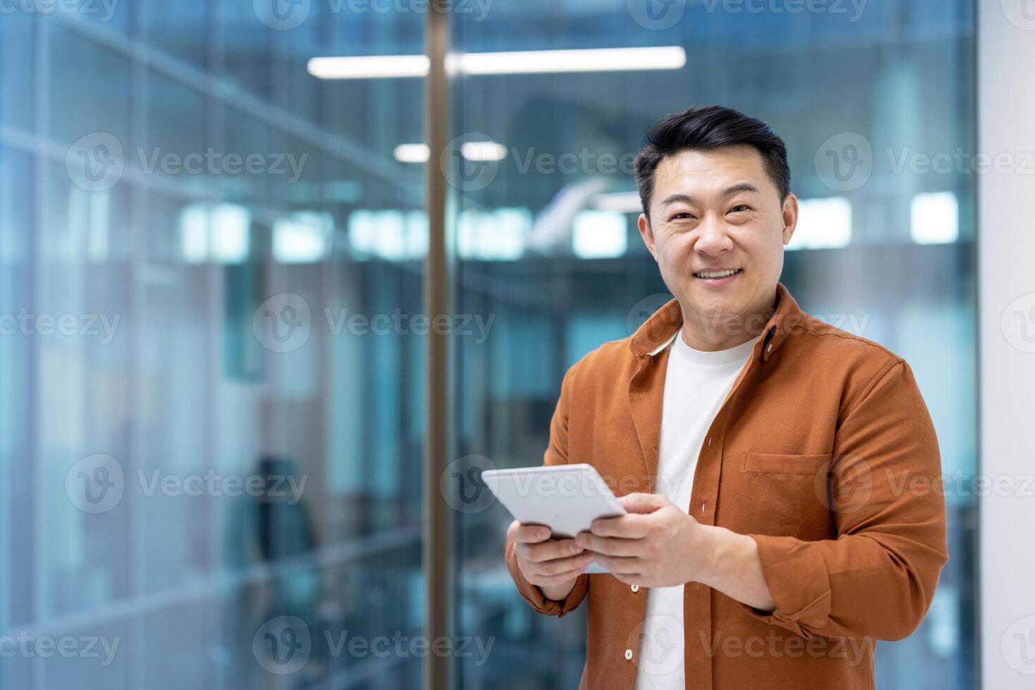 Portrait of an Asian man inside an office at his workplace, a man looks at the camera, smiles while holding a tablet computer. Mature businessman in casual clothes stands near the window. photo