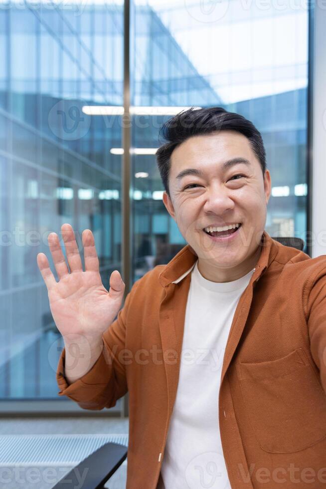 call, Asian man looking into phone camera and talking to colleagues using smartphone app. Businessman at workplace inside office. photo