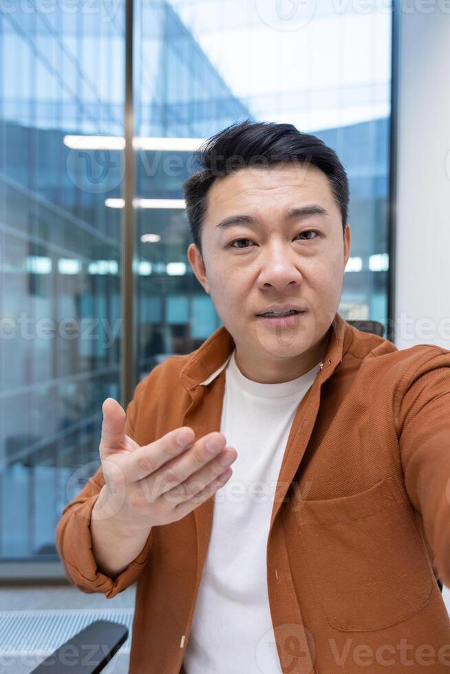 Asian mature businessman engaging in a call, looking at the camera talking. Located indoors with a professional ambiance, conducting a serious and compelling conversation for communication photo