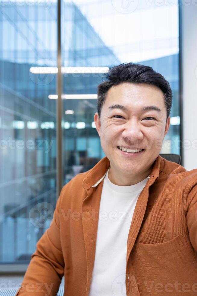 An Asian mature businessman takes a selfie call with his phone, smiling confidently in a modern office environment, expressing professionalism and a positive demeanor. photo