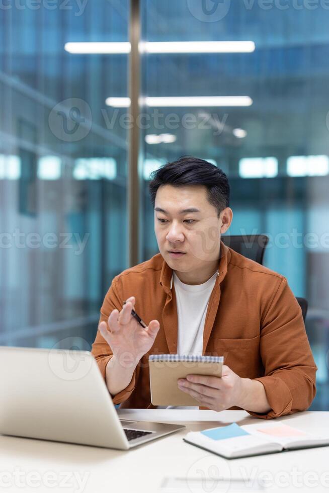 Asian man mature businessman engaged in remote work, taking notes, and communicating online in a modern workspace. Represents productivity and digital work environments. photo