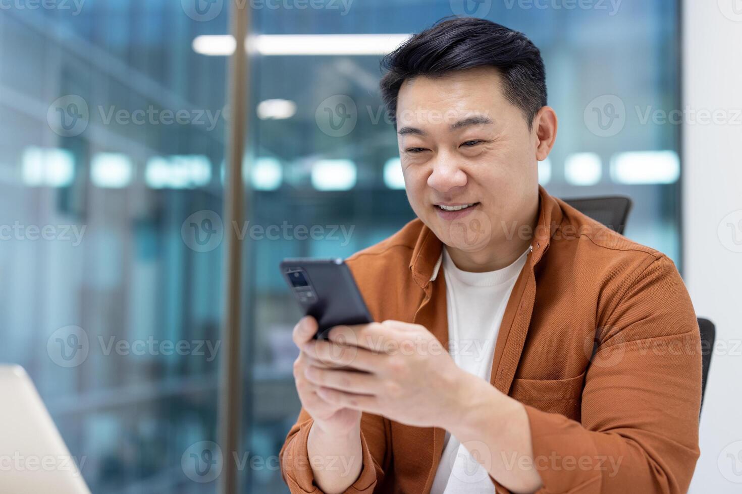 Close-up of successful Asian man using phone, smiling man typing text message, satisfied businessman in casual shirt inside office. photo