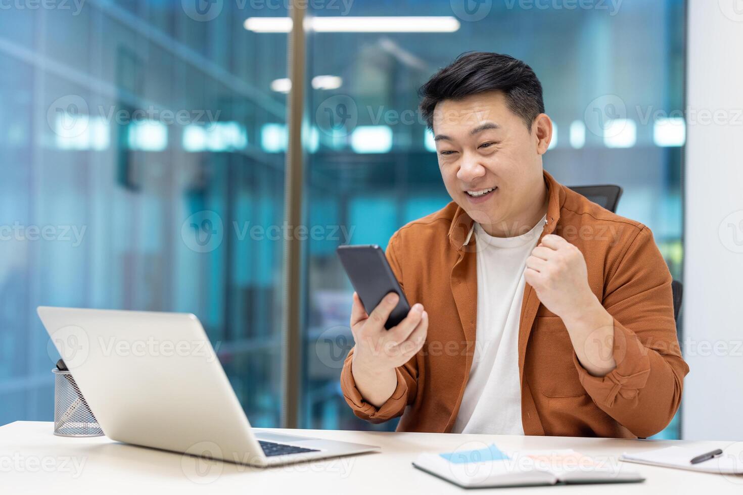 Asian businessman showing excitement looking at phone at office desk. Celebrating success in professional environment with modern tech tools around. Positive energy and motivation. photo