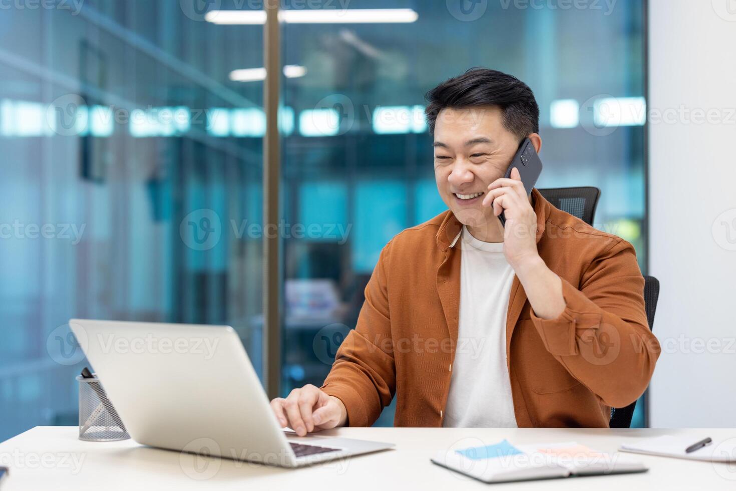 Asian businessman smiling while multitasking with phone and laptop in modern office setting, demonstrating effective communication and productivity. Concept of technology in business environment. photo