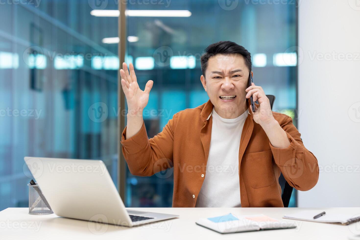 Asian businessman expressing frustration during business call in modern office. Man seated at desk with laptop, using phone, gesturing in conversation, showcasing emotion and communication. photo