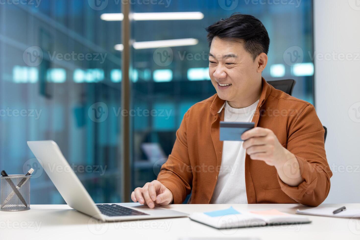 Asian businessman smiling while using credit card and laptop in modern office setting. Confident entrepreneur engaging in online shopping. Business, technology, e-commerce, finance concept. photo