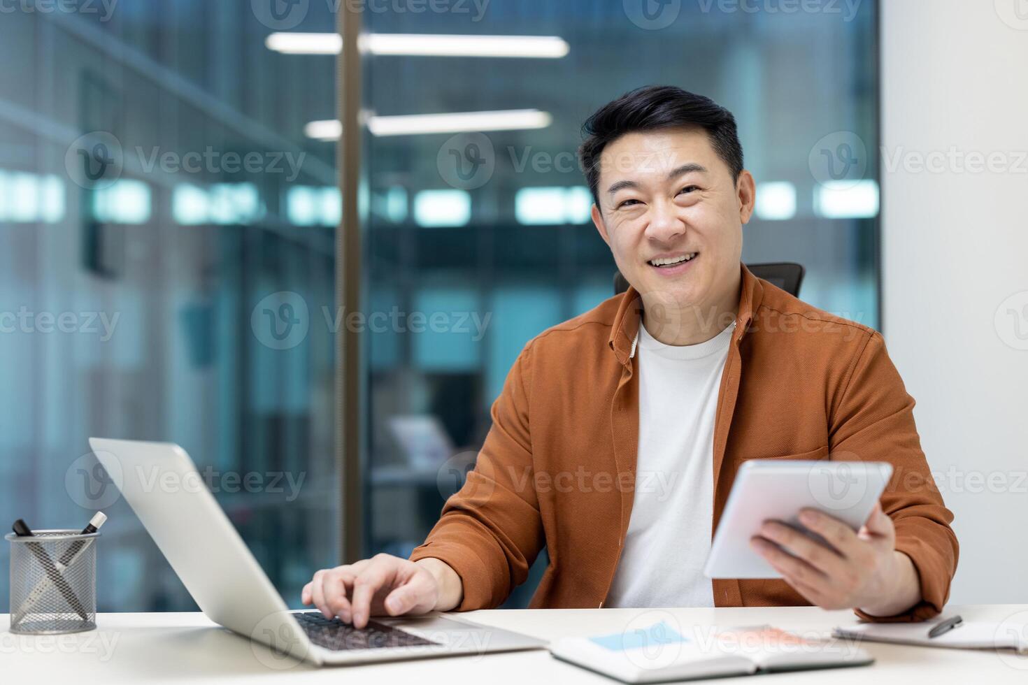 Asian man mature businessman working in an office using a tablet and laptop, smiling confidently. The atmosphere reflects productivity, success, and a positive attitude. photo