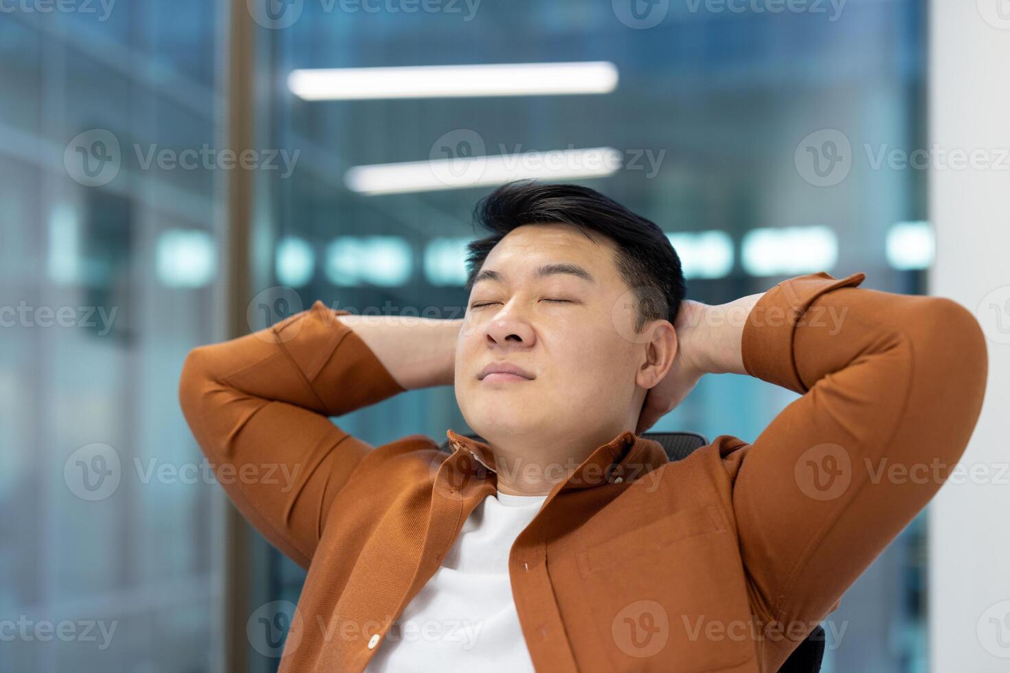 Asian man resting and relaxing close up. Businessman working inside office close up with eyes closed and hands behind head. photo