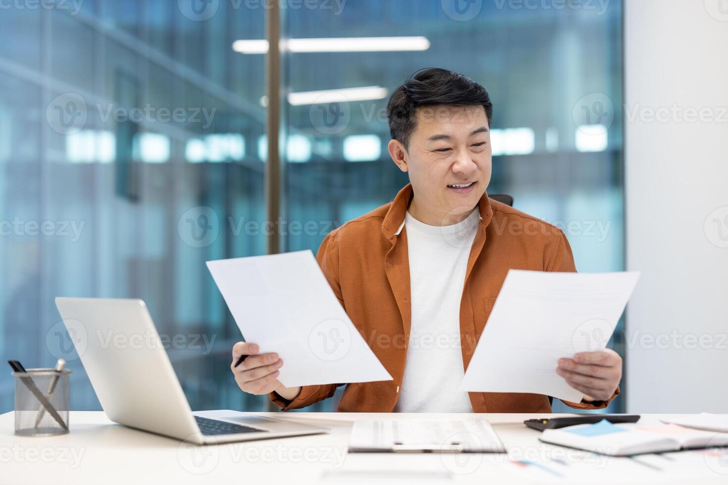 Successful Asian financier checking investment reports. Happy satisfied man smiling holding documents and papers in hands. Businessman at workplace inside office doing paperwork. photo