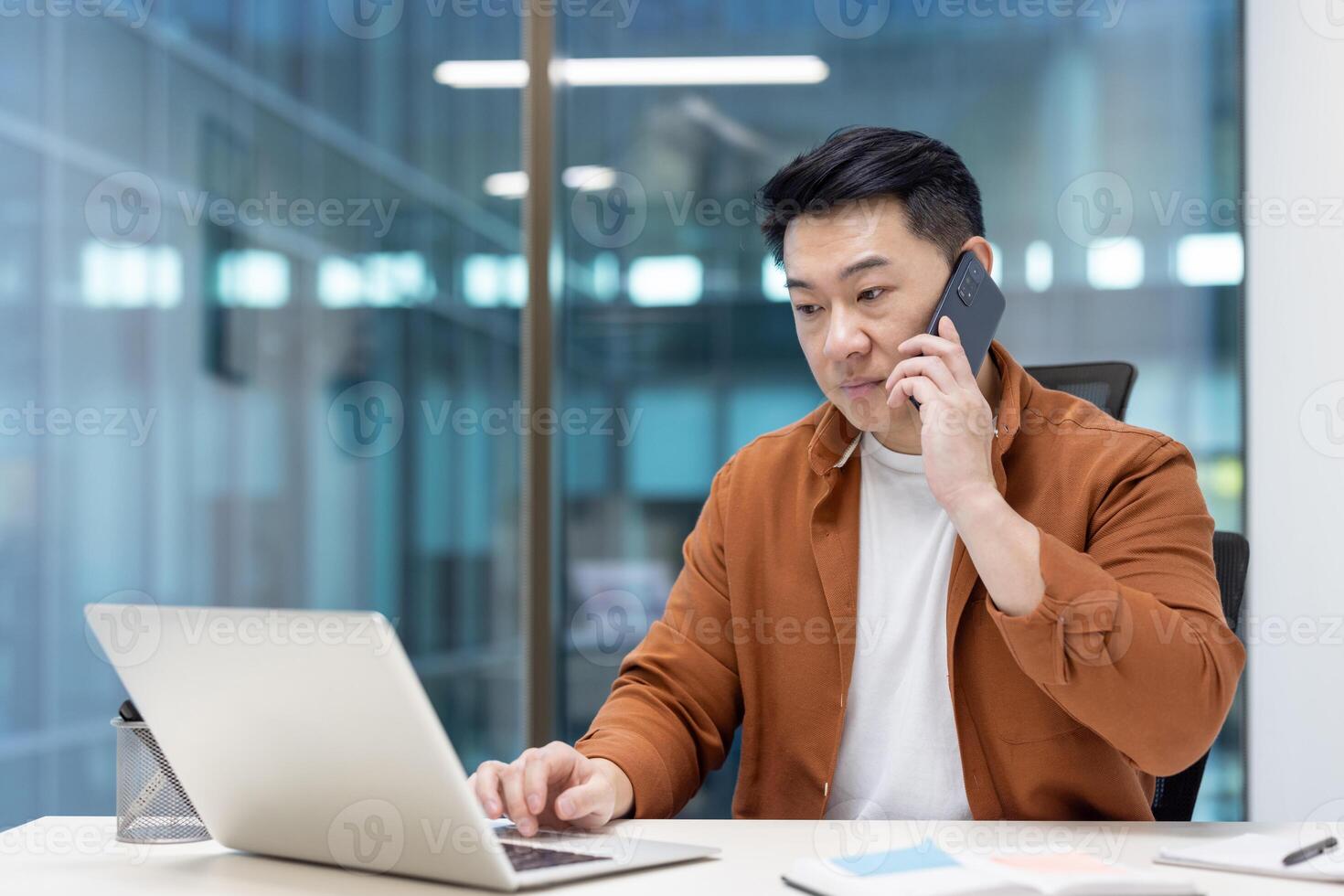Serious and focused successful Asian businessman talking on mobile phone. Male office worker at workplace inside office using laptop. photo
