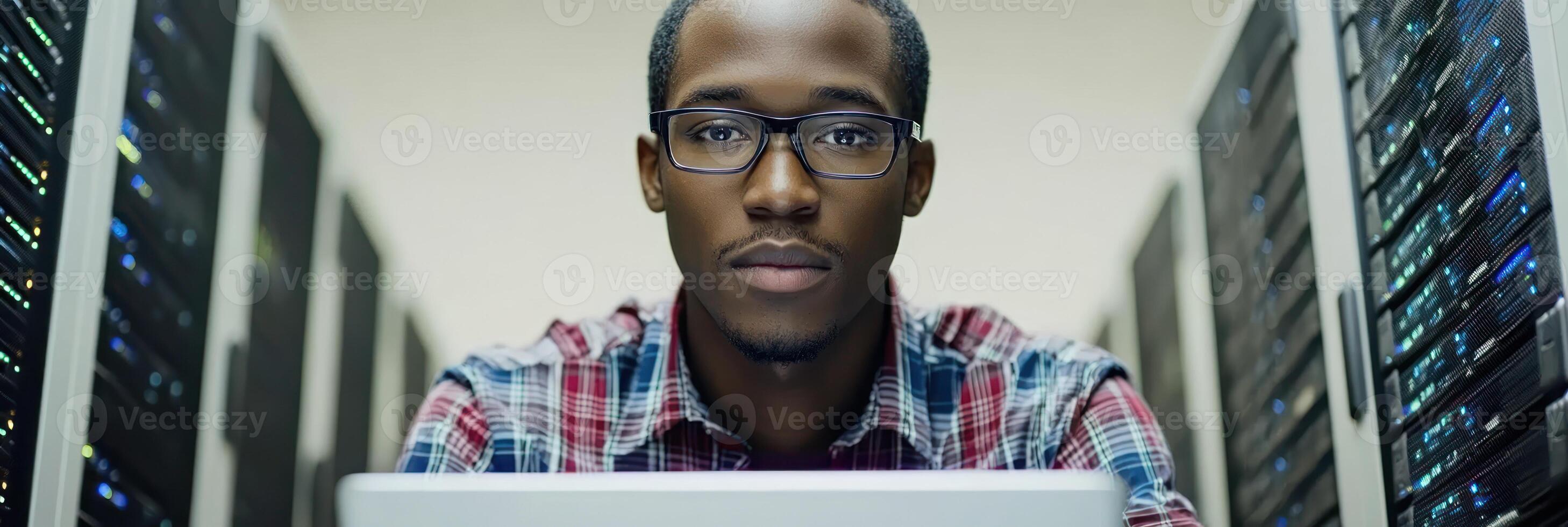 Focused young man operates laptop in server room filled with data servers and equipment photo