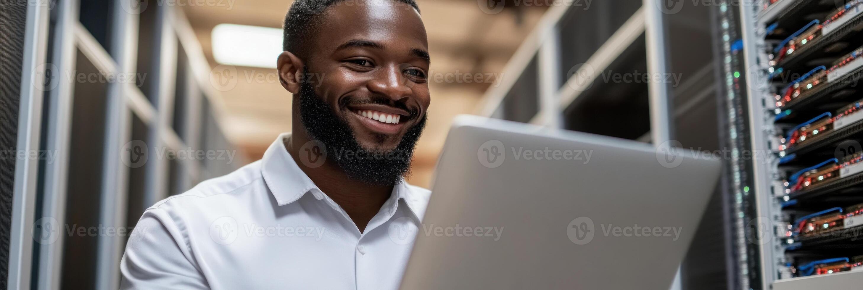 Man smiles while using a laptop in a busy data center with numerous server racks around photo