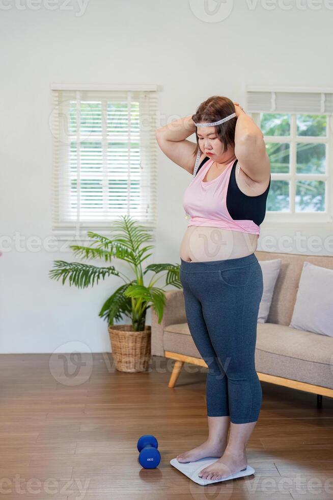 Overweight woman standing on a scale, preparing to workout with dumbbells at home, focusing on weight loss photo