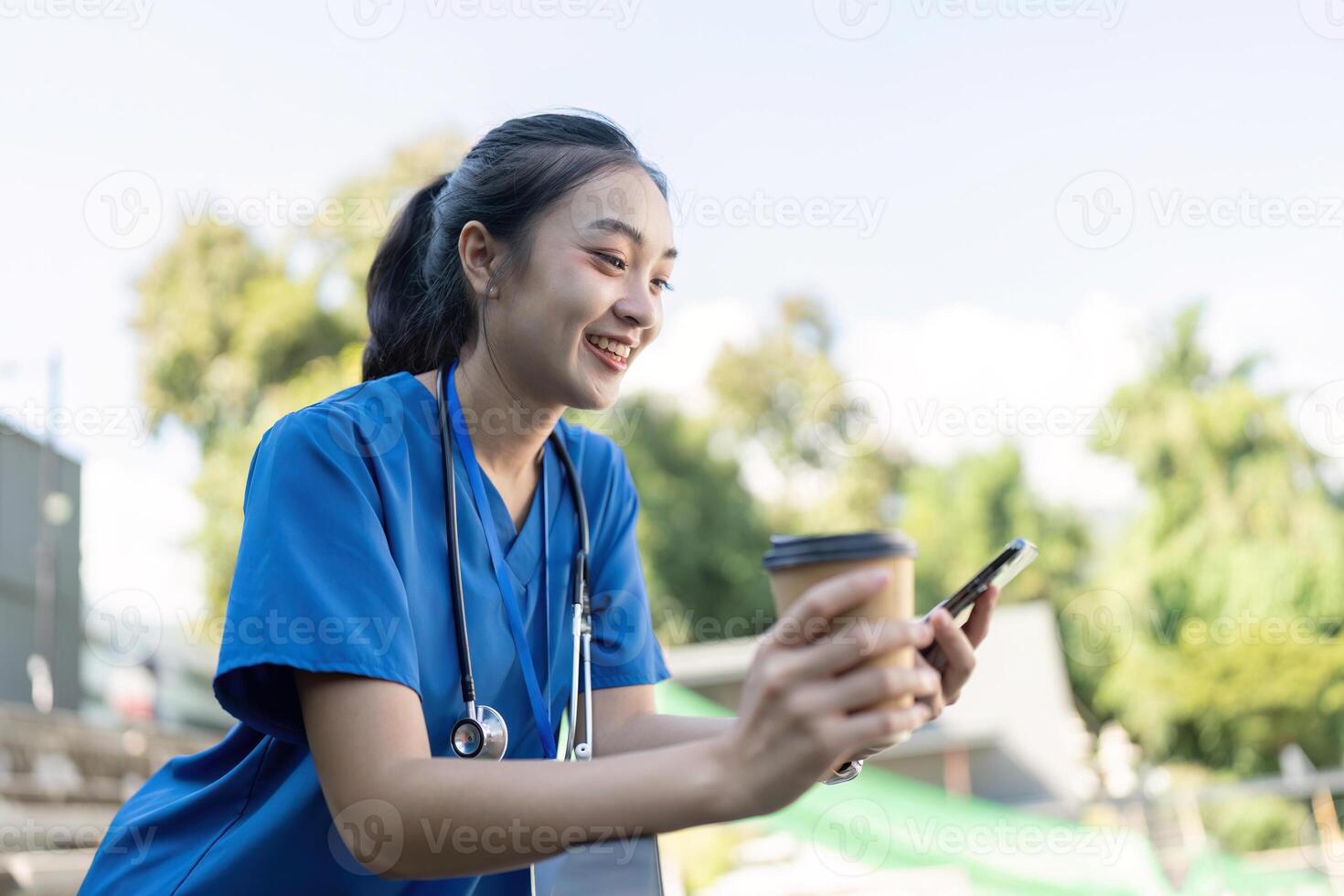 Nurse smiling while holding a coffee cup and smartphone, enjoying a sunny break in a healthcare environment photo