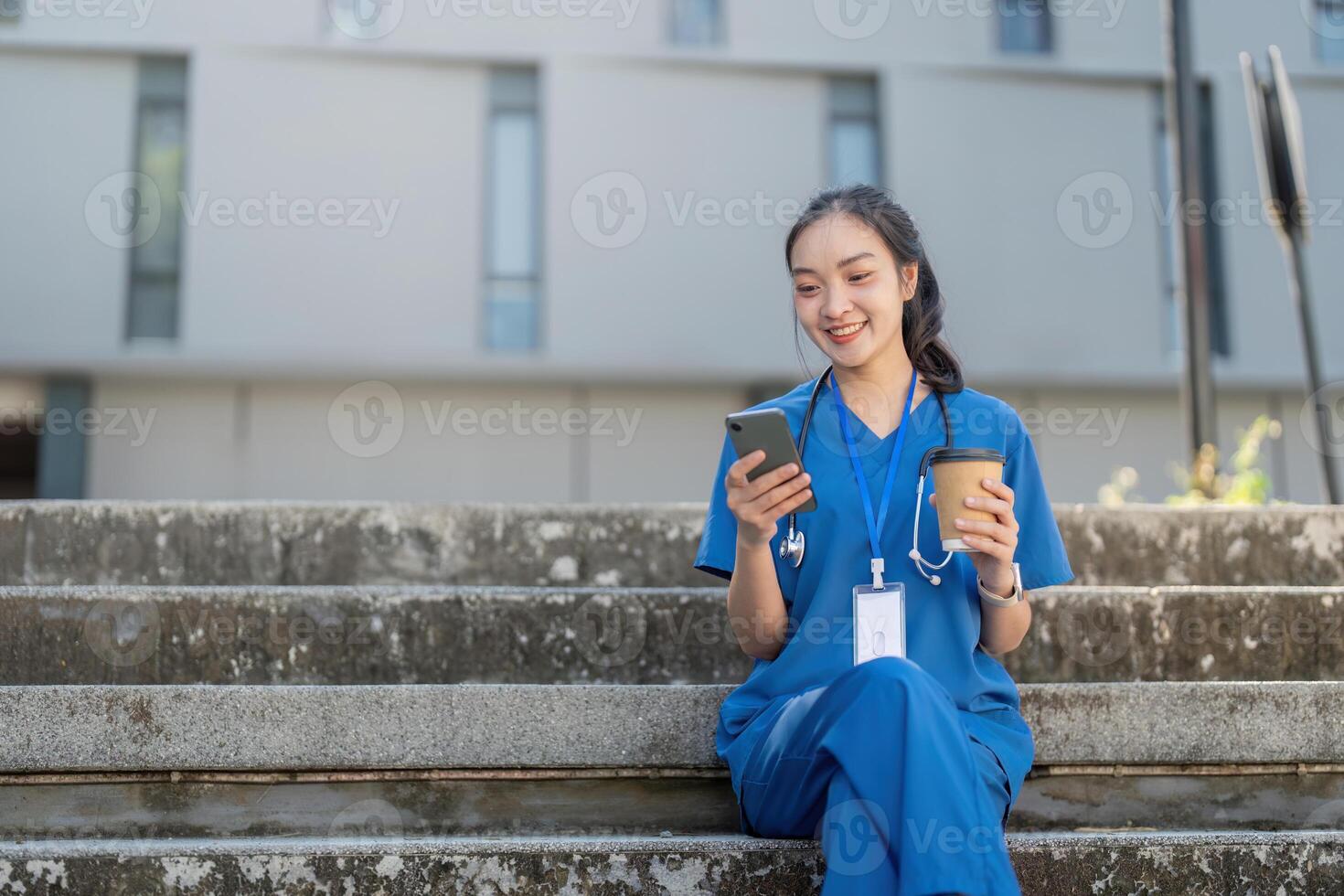 Nurse sitting on steps, smiling while using her smartphone and holding a coffee cup in a sunny healthcare environment photo
