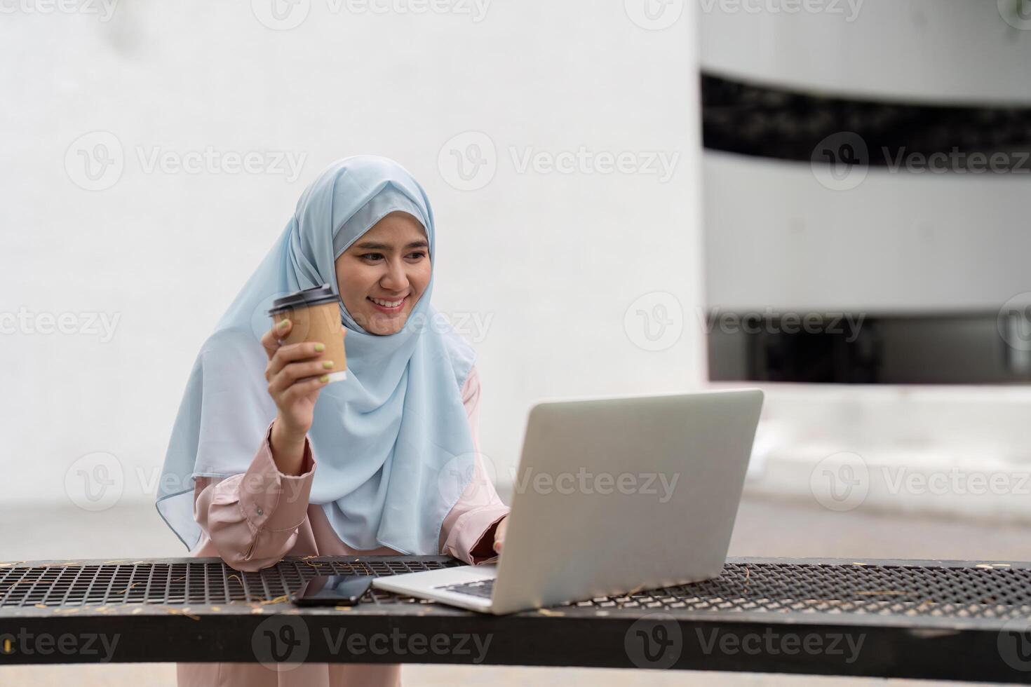 Muslim woman smiling while holding coffee and working on a laptop in a business setting. photo