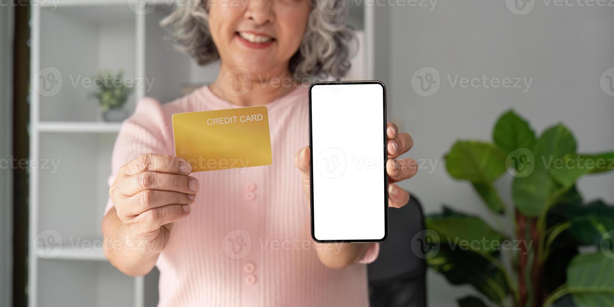 Elderly woman smiling and holding a credit card next to a smartphone with a blank screen. photo