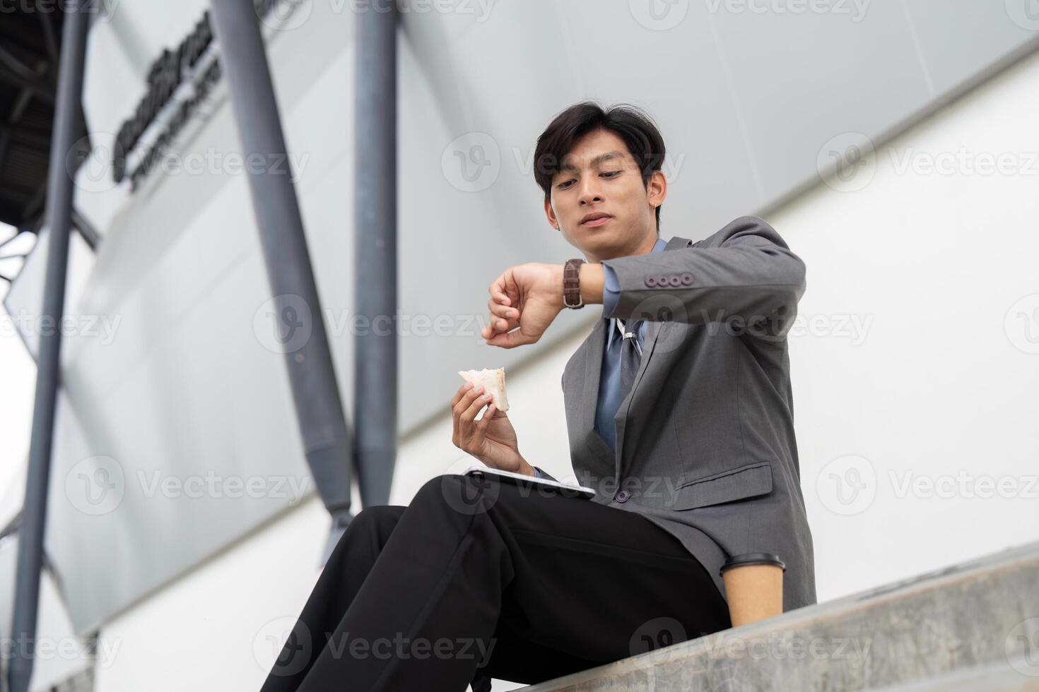Young Asian businessman checking the time while enjoying a snack in a modern outdoor business setting. photo