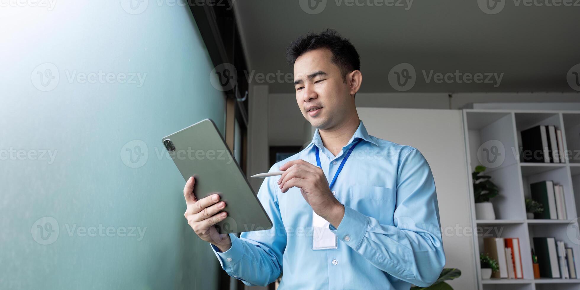 Focused Asian businessman using a tablet for planning, standing by a window in a modern office environment. photo