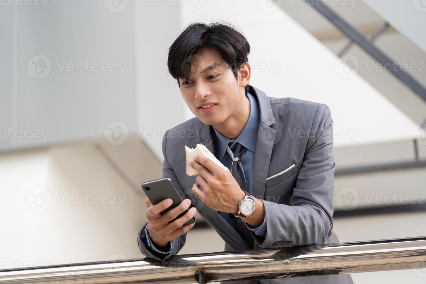 Young Asian businessman enjoying a snack while checking his phone in a modern office setting. photo