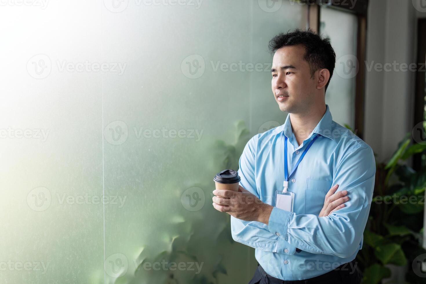 Contemplative Asian businessman holding a coffee cup, standing with arms crossed by a frosted glass window in a modern office. photo