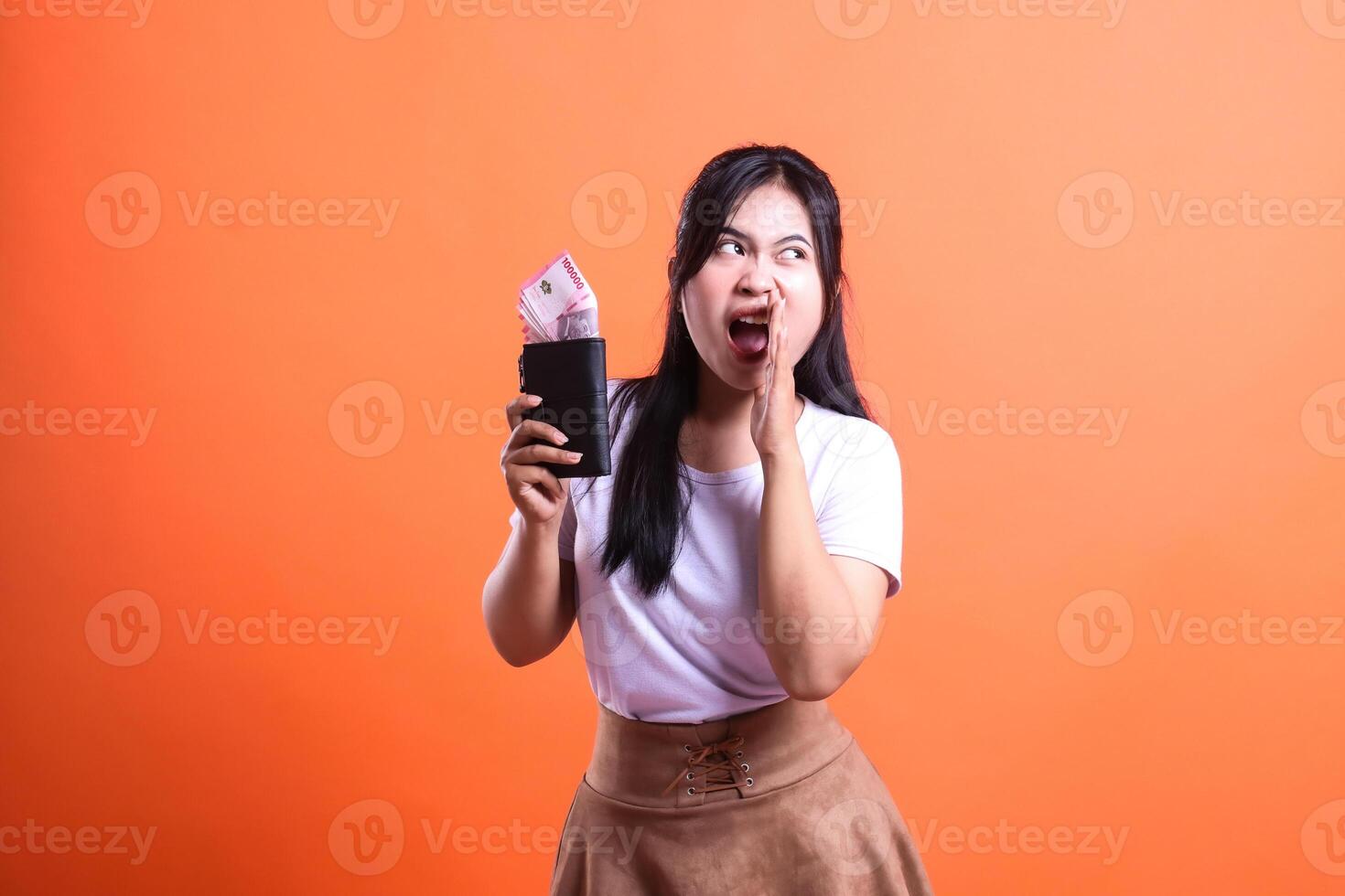 A woman holds a black wallet full of Rupiah banknotes, with a surprised expression while whispering. She is wearing a white shirt and brown skirt, isolated on an orange background. photo